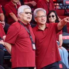 Two people wearing red shirts and ID lanyards stand and converse in a group setting with blurred audience chairs labeled with names in the background.