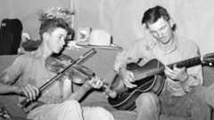 Black and white photo of two people with short, greased-back hair sitting on a couch and playing a violin and a guitar.