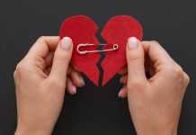 Hands holding a broken red heart patched together with a safety pin against a black background.