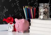 A pink piggy bank, a jar of coins, books, and colorful pencils on a desk against a chalkboard background