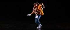 Justin Peck lifts Patricia Delgado as he moves toward stage right. Delgado leans forward, toward where they are going, one leg extended front and the other bent, her outside arm raised at the elbow. Peck wears jeans and white sneakers. Delgado wears a mustard yellow dress and white sneakers.