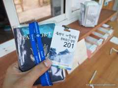 A hand on the left of the frame holding a passport and a permit with a blue lanyard. In the background is a wooden desk with office supplies in soft focus.
