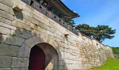 Stone block wall with an arched doorway, at Seoul fortress, a roof visible in the background, and trees and blue sky behind.