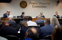 Four people sit at a head table in a meeting room under a sign that says "Public Utilities Commission, with an audience in the foreground.