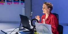 A woman in a red shirt is sitting at a desk, smiling and gesturing animatedly while speaking into a microphone. She is in front of a laptop with a circular light ring, and there are papers and cables on the desk. The background is blurred with photos and Post-it notes on the wall.