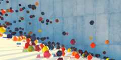 A surreal scene features colorful, multi-faceted geometric shapes floating in the air and resting on white steps in front of a large, textured blue wall. The shapes vary in color, including reds, yellows, oranges, blues, and greens.