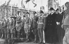 Adolf Hitler stands at the head of a large group of uniformed soldiers and officials. He raises his arm in a Nazi salute towards a parade occurring out of frame.