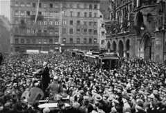 A large crowd fills the city square. A singular individual stands up on a makeshift dais and talks to the crowd. A streetcar can be seen in the background, surrounded by people.