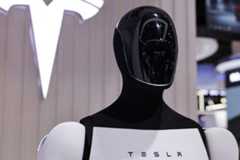Robô humanoide preto e branco com rosto brilhante e sem traços faciais, exibido em estande com logo da Tesla ao fundo.