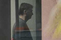 Homem de perfil, com cabelo curto e escuro, está em ambiente interno com iluminação baixa. Ele veste camiseta escura e está próximo a uma parede clara, com reflexos e sombras visíveis na imagem.