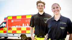 Eine junge Notfallsanitäterin und ein junger Notfallsanitäter stehen in Rettungsdienstbekleidung vor einem Rettungswagen.
