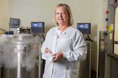 A woman in a white lab coat stands in a lab, holding a white box, with cryogenic storage tanks behind her.