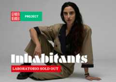 Luna Cenere è seduta a terra in posa rilassata, ha lunghi capelli scuri, indossa una tuta beige con dettagli blu scuro e anfibi neri. Sullo sfondo grigio chiaro, in alto a sinistra compare il logo di Gender Bender e la scritta 'PROJECT' su sfondo verde.