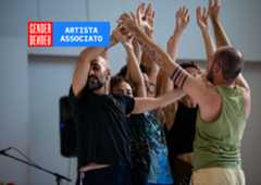 Un gruppo di performer in scena durante una prova o una performance: sono molto vicini tra loro, con le braccia sollevate e intrecciate, creando una composizione corale di corpi ed espressioni intense