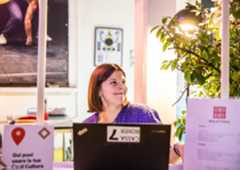 Donna sorridente dietro al banco biglietteria di un teatro, seduta al computer, con luce calda e pianta decorativa accanto.