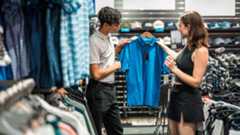Young diverse couple, male and female, excitedly browsing golf apparel in a sports store. The man holds a blue golf shirt, while the woman observes. Both are casually dressed in summer outfits, exploring options amidst golf gear displays.