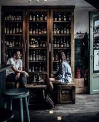 two people sitting at a table in front of a shelf full of bottles and glasses