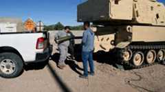Personnel load an artillery round beside a tracked armored vehicle at a training site, with equipment and support vehicles nearby.