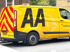 A yellow van with the letters "AA" prominently displayed on its rear side, parked on the side of the road. The van features a distinctive red and yellow reflective strip along its side and a black border around the windows.