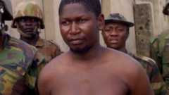 Mohammed Yusuf, bare-chested and with a bandage on his arm, surrounded by soldiers