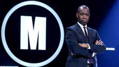 Masterming host Clive Myrie stands with his arms folded on the Mastermind set. He wears a dark suit and there's a large white glowing M in a circle on the wall behind him.