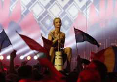 Eine Frau sitzt auf einer hölzernen Buttermelkanne, umgeben von jubelnden Menschen mit Flaggen.