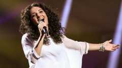 Eine Sängerin mit lockigem Haar singt in ein Mikrofon auf einer Bühne.