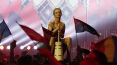 Eine Frau in Tracht sitzt auf einem Butterfass, umgeben von Fahnen schwenkenden Zuschauern.