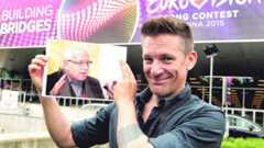 Ein Mann hält ein Foto von Marcel Reich-Ranicki vor einem Eurovision Song Contest-Banner.