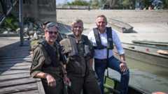 Drei Männer in Schwimmwesten posieren auf einem Steg neben mehreren Booten.