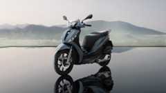 Ein graublauer Motorroller steht vor einer Bergkulisse und spiegelt sich im Wasser. Ein graublauer Motorroller steht vor einer Bergkulisse und spiegelt sich im Wasser.