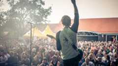 Ein Musiker mit Gitarre steht auf einer Bühne vor einer großen, jubelnden Menschenmenge im Freien.