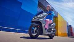 46-217900283 Eine Frau fährt mit einem grauen Honda-Motorroller vor einem farbenfrohen Gebäude.