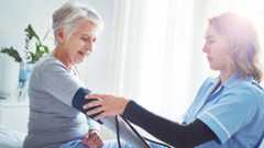 Ältere Frau sitzt, während eine Pflegekraft in blauer Arbeitskleidung ihren Blutdruck misst.