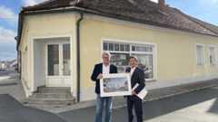 Zwei Männer stehen vor einem gelben Haus und halten gemeinsam eine Zeichnung eines modernen Gebäudes.