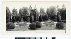 Symmetrisch angelegter Garten mit runden Sträuchern, Blumenbeeten, Denkmälern und Bäumen im Hintergrund.