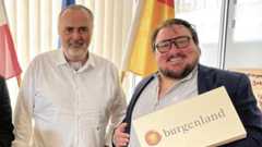 Zwei Männer stehen nebeneinander, einer hält eine Geschenkbox mit der Aufschrift „Burgenland“, im Hintergrund sind Flaggen zu sehen.