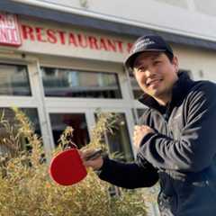 Ein lächelnder Mann mit einem Tischtennisschläger vor einem Restaurant namens „Ding 1900“.