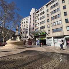 Ein Platz mit einem Brunnen und angrenzenden Gebäuden an einem sonnigen Tag.