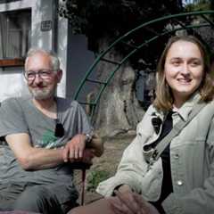 Ein Mann und eine junge Frau sitzen draußen vor einem Gebäude mit einem Fass im Hintergrund.