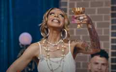 Person joyfully raising a gold goblet, wearing a sleeveless dress with layered necklaces and hoop earrings