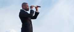 A person in a suit looking through a telescope against a blue sky, symbolizing vision or looking towards the future.