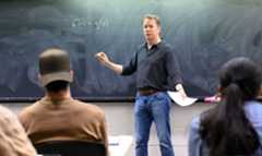 Faculty (Sean Caroll) in front of a chalk board teaching a class of undergraduate students.