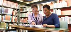 Two women looking at an open book while surrounded by shelves of books