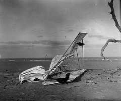 Photo shows a glider folded in a heap on the beach with the ocean beyond.