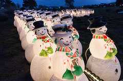 Two long rows of smiling snowmen figures, lit from within, are each wearing a black hat, green gloves, and red and green scarf while holding a candy cane.