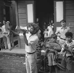 A man in the foreground stands in the street and speaks through a paper megaphone. Two boys near him hold a young goat and live chicken. Men and children watch from the sidewalk.