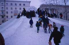 Children and some adults are walking up the middle of a snow-covered hill in a city and sliding down the side of the hill. An icy arch in the background has '1999' on it.