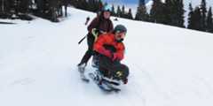 Sean Marihugh smiles as he slides down a ski slope in a sit-ski with a partner on skis holding onto the back of his sit-ski.