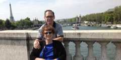 Barbara and Jim Twardowski pose on a bridge over the Seine in Paris with the Eiffel Tower visible in the background. Barbara is sitting in a wheelchair and Jim is standing behind her.
